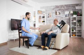 Male nurse on a chair in a nursing home talking with elderly age man while he is sitting on couch.